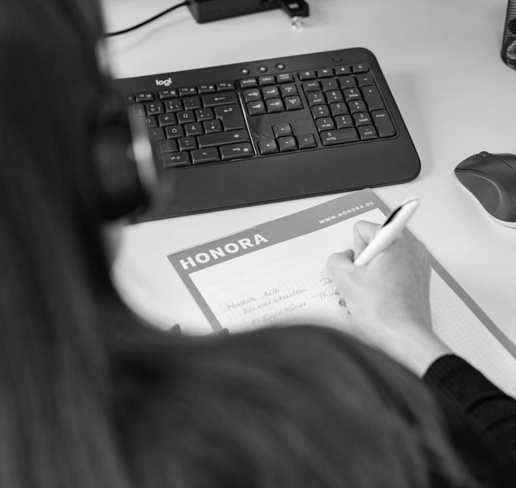 Symbolbild, ein Honora Mitarbeiter arbeitet mit Headset an seinem Schreibtisch und schreibt etwas auf einen Block